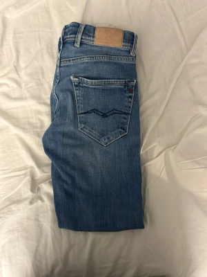 Blå raka jeans från Replay - Säljer ett par klassiska blå jeans från Replay med raka ben och femficksmodell. Jeansen har snygga slitningar framtill och en läderpatch med logga bak i midjan. Materialet är mjukt denim som sitter skönt hela dagen.