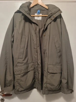 Olivgrön parkas från Peak Performance - Snygg olivgrön parkas från Peak Performance med huva, dragkedja och tryckknappar. Jackan har två stora fickor med lock och två snedställda bröstfickor. Tillverkad i slitstarkt material, perfekt för kyliga dagar. Stilsäker och funktionell design.