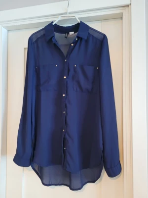 Mörkblå skjorta från H&M - Stilren mörkblå skjorta från H&M med transparent känsla. Skjortan har långa ärmar, två bröstfickor och guldfärgade knappar framtill och vid ärmslut. Perfekt för dig som gillar en enkel men snygg look.