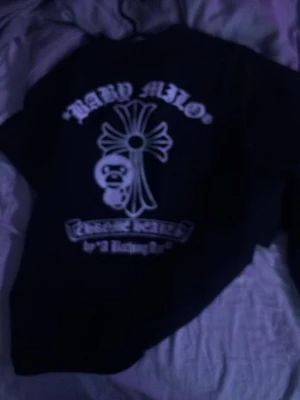 Svart Chrome Hearts x Bape T-shirt - Säljer en svart t-shirt med stora vita tryck från Chrome Hearts och Bape. Framsidan har klassiska Chrome Hearts-loggan med Bape-ansiktet i mitten, och baksidan har Baby Milo och ett kors. Perfekt för dig som gillar streetwear och unika samarbeten.