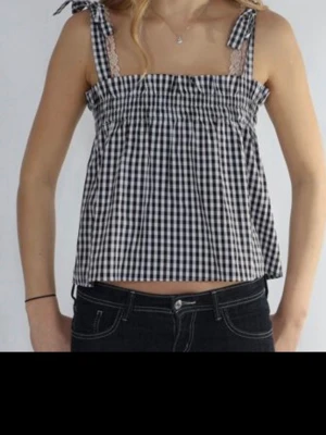 Rutig svartvit linnetopp med rosetter - Söt linnetopp med svartvitt rutigt mönster och knytband på axlarna. Toppen har en luftig passform och spetsdetaljer vid axelbanden. Perfekt för varma dagar och ger en chill vibe till din outfit.