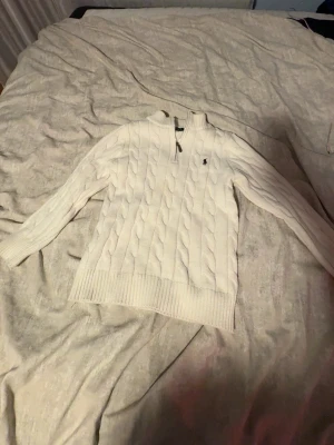 Vit kabelstickad tröja Polo Ralph Lauren - Kabelstickad vit tröja från Polo Ralph Lauren med halv dragkedja och liten broderad logga på bröstet. Tröjan har ribbade muddar vid ärmslut och nederkant samt hög krage. Perfekt för lager på lager och klassisk stil.