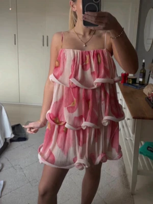 Rosa blommig volangklänning - Supersöt kort klänning med rosa och vita blommor, volangdetaljer och tunna axelband. Klänningen har en luftig passform Materialet känns lätt och flowy