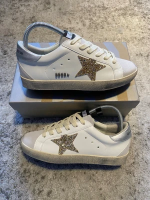 Golden Goose vita sneakers med glitterstjärna - Säljer ett par vita Golden Goose sneakers med en stor glittrig guldstjärna på sidan. Skorna har beige detaljer vid tån och hälen, samt en slitstark sula i grått. Klassisk låg modell med vita skosnören och perforerade hål på sidan.
