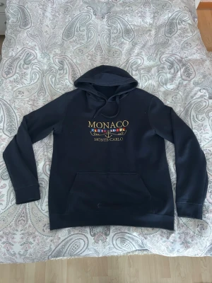 Marinblå Monaco Monte-Carlo hoodie - Snygg marinblå hoodie med huva och dragsnören. På bröstet står det 'MONACO MONTE-CARLO' i gul text med broderade flaggor och ett ankare. Går endast att köpa lokalt i Monaco, nypris 1700 mitt pris 399. Perfekt för dig som gillar stilren och nautisk design.