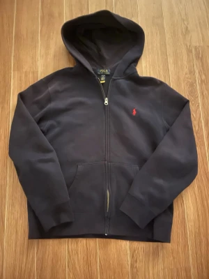 Mörkblå hoodie från Polo Ralph Lauren - Snygg mörkblå hoodie med dragkedja från Polo Ralph Lauren. Klassisk design med huva, fickor framtill och den ikoniska röda logon på bröstet. Perfekt för dig som gillar stilrena och bekväma plagg. Materialet är mjukt och känns riktigt skönt mot huden. Storlek XS, L(14-16). Bara hör av er vid funderingar! 😃