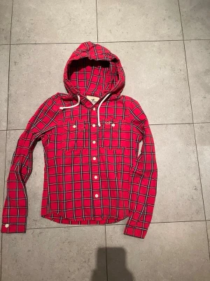 Hollister tröja  - Säljer en röd hoodie från Hollister med svart och vit rutmönster. Hoodien har knappar framtill, två bröstfickor med lock och vita dragsnören i huvan. Tillverkad i mjukt tyg, perfekt för en avslappnad och trendig stil.