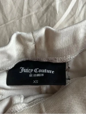 Vita mjukisbyxor Juicy Couture XS - Vita mjukisbyxor från Juicy Couture i storlek XS. Byxorna har snörning i midjan, raka ben och bakfickor med knappdetalj. Bakdelen av ena knappen fattas! Tillverkade i mjukt frottématerial som ger en chill och lyxig känsla.