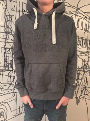 Superdry hoodie - Mörkgrå hoodie från Superdry med broderad logga på bröstet och japansk text. Stora vita dragsnören i huvan och klassisk känguruficka framtill. Ribbstickade muddar vid ärmslut och nederkant. Perfekt för en chill och avslappnad stil.