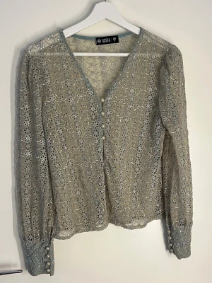 Blommig spetsblus från CUTE SAINT - Snygg transparent spetsblus i beige med blommigt mönster och pärlknappar framtill. V-ringad med långa ärmar och dekorativa knappar vid ärmsluten. Passar xs/S