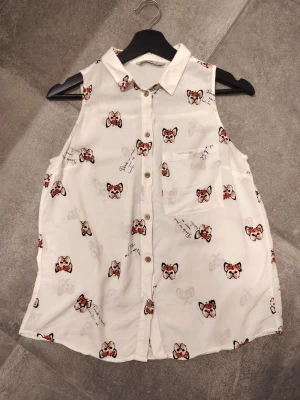 Vit ärmlös blus med söt mönster - Söt vit blus med krage och knäppning framtill. Blusen är ärmlös och har ett lekfullt tryck med små hundansikten med blommor i rött och svart samt diskreta textdetaljer. En bröstficka på vänster sida. Perfekt till ett par jeans. Den är i jättefint skick då den knappt har använts, vart för liten. Köptes utomlands, storlek S