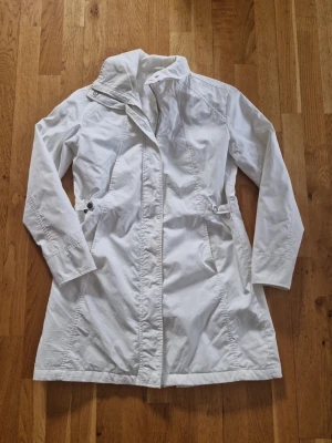 Vit trenchcoat från Colours Woman - Stilren vit trenchcoat från Colours Woman med hög krage och knäppning framtill. Jackan har två fickor med knappdetaljer i midjan och en figurnära passform. Perfekt för dig som vill ha en klassisk och enkel look. Materialet känns lätt och smidigt.