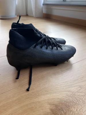 Svarta Nike fotbollsskor - Säljer ett par svarta Nike fotbollsskor med högt skaft och snörning. Skorna har en modern, strömlinjeformad design och är tillverkade i syntetmaterial med mönstrad yta för bättre bollkontroll. Dobbarna är anpassade för spel på gräsplan.