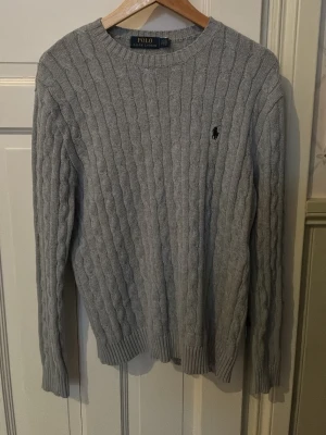 Grå kabelstickad tröja Polo Ralph Lauren - Kabelstickad grå tröja från Polo Ralph Lauren med rund halsringning och diskret broderad logga på bröstet. Tröjan har långa ärmar och ribbade muddar vid ärmslut och nederkant. Klassisk och stilren design i mjukt material. Säljer den enbart för den blev för liten, mycket bra kvalitet 