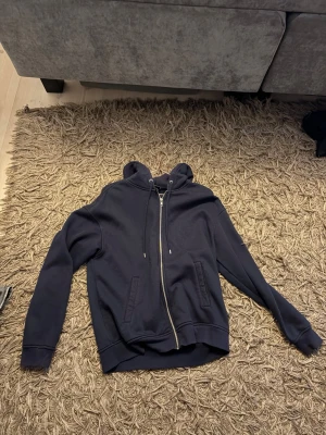 Mörkblå hoodie med dragkedja - Snygg mörkblå hoodie med dragkedja framtill och två fickor. Klassisk modell med huva och dragsnören. Perfekt för en avslappnad och stilren look. Passar till det mesta och är enkel att matcha med andra plagg.