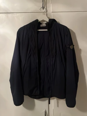 Mörkblå vindjacka från Stone Island - Snygg mörkblå vindjacka från Stone Island med huva och klassisk patch på vänster ärm. Jackan har en minimalistisk design, dragkedja framtill och är tillverkad i ett lätt, vindtätt material som passar perfekt för blåsiga dagar. 