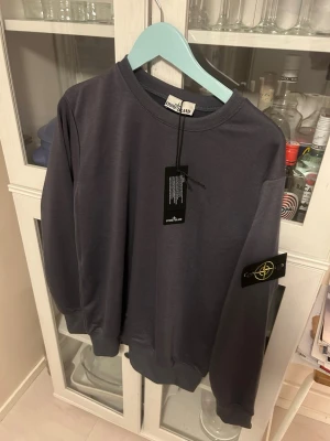 Mörkblå sweatshirt från Stone Island - Säljer en mörkblå sweatshirt från Stone Island med den klassiska loggan på vänster ärm. Tröjan har rund halsringning, ribbade muddar och är gjord i mjukt bomullsmaterial. Perfekt för dig som gillar streetwear och vill ha en clean look.
