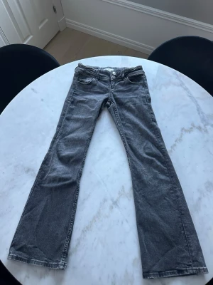 Grå bootcut jeans med fickdetaljer - Snygga grå bootcut jeans med klassiska fem fickor och knappar på bakfickorna. Jeansen har låg midja och utsvängda ben.