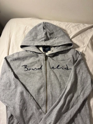 Grå zip hoodie från Bondelid - Snygg grå hoodie från Bondelid med dragkedja framtill och huva. Tröjan har ett broderat Bondelid-tryck över bröstet och är gjord i mjukt bomullsmaterial. Perfekt för en avslappnad och stilren look.