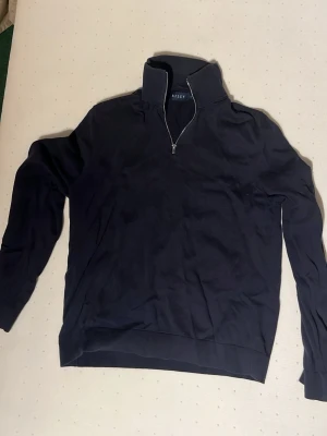 Svart halvzip tröja från Riley - Stilren svart långärmad tröja från Riley med halv dragkedja och hög krage. Perfekt för lager på lager och enkel att matcha med olika outfits. Tillverkad i mjukt material för skön känsla hela dagen.