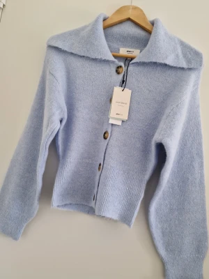 Ljusblå stickad kofta från Gina Tricot - Superfin ljusblå kofta från Gina Tricot i mjuk ullblandning. Koftan har stora bruna knappar framtill, bred krage och ribbade muddar vid ärmslut och midja. Perfekt för lager på lager och ger en mysig look. Storlek XS. Ny med prislapp.