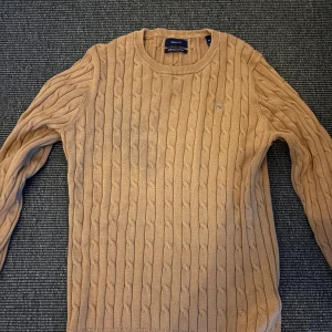 Beige kabelstickad tröja från Gant - Säljer en beige kabelstickad tröja från Gant med rund halsringning och diskret logga på bröstet. Tröjan har långa ärmar och ribbade muddar vid ärmslut och nederkant. Perfekt för dig som gillar klassisk och stilren design.