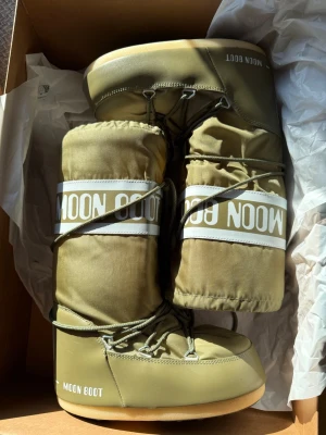 Olivgröna Moon Boot snow boots - Säljer ett par olivgröna Moon Boot snow boots med vit logga och snörning runt skaftet. De har en klassisk, vadderad form och är perfekta för kalla dagar. Yttermaterialet är syntet och sulan är i gummi. Riktigt ikoniska vinterboots för dig som vill sticka ut.