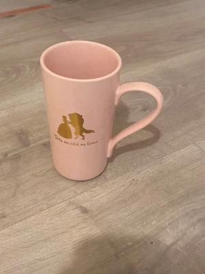 Rosa Beauty and the Beast mugg - Säljer en ljusrosa mugg med motiv från Beauty and the Beast. Muggen har guldfärgad text och siluett av Belle och Odjuret samt citatet 'Tale as old as time'. Perfekt för Disneyfans som vill ha något unikt till samlingen.