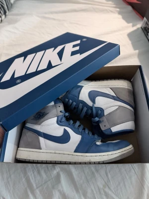 Nike Air Jordan 1 High OG blå/vit - Snygga Nike Air Jordan 1 High OG sneakers i blått, vitt och grått. Skorna har klassisk hög siluett, blå swoosh och detaljer, samt grå mocka på hälen. Ovandelen är i läder och mocka, med Air Jordan-logga på sidan. Perfekta för dig som gillar streetwear och ikoniska sneakers.