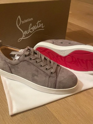 Gråa Christian Louboutin sneakers - Snygga gråa sneakers från Christian Louboutin i mocka med klassisk röd sula och vita kanter. Skorna har lågt skaft, rund tå och matchande grå skosnören. Perfekta för dig som vill sticka ut med exklusiv design och ikoniska detaljer.