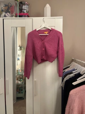 Rosa cropped stickad kofta - Säljer en rosa cropped stickad kofta med v-ringning och stora knappar framtill. Koftan har en chunky stickning och långa ärmar, perfekt för att lägga till färg i din outfit. Passar dig som gillar trendiga och bekväma plagg.