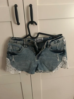 Blå jeansshorts med spetsdetaljer - Säljer ett par blå jeansshorts från Inside med slitningar och snygga vita spetsdetaljer längs kanterna. Shortsen har klassisk femficksdesign och rå kant nedtill för en avslappnad look. Perfekta för varma dagar!