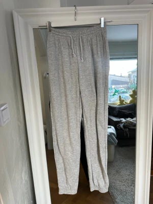 Grå mjukisbyxor från Gina Tricot - Säljer ett par ljusgrå mjukisbyxor från Gina Tricot med dragsko i midjan och resår vid bensluten. Byxorna är tillverkade i mjukt sweatshirtmaterial och har en avslappnad passform som är perfekt för chill dagar.