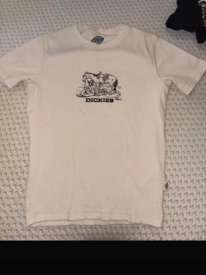 Vit ribbad t-shirt från Dickies - Säljer en vit ribbad t-shirt från Dickies med svart tryck framtill som föreställer en cowboy och en häst. Klassisk rund hals och korta ärmar. Perfekt för dig som gillar streetwear och vill ha något unikt från Dickies.