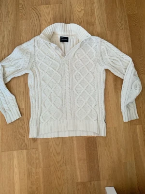 Vit kabelstickad tröja Peak Performance - Snygg vit kabelstickad tröja från Peak Performance med krage och två knappar vid halsen. Tröjan har ett klassiskt mönster framtill, ribbade muddar och är tillverkad i ett mjukt, tjockt material. Perfekt för kalla dagar och enkel att matcha.