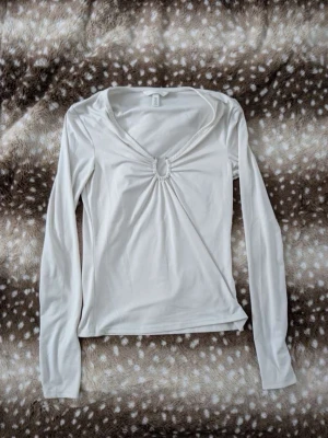 Vit långärmad topp med ringdetalj - Trendig vit långärmad topp med v-ringning och snygg ringdetalj framtill som ger en cool twist. Tillverkad i mjukt material som känns skönt mot huden. Perfekt att styla med jeans eller kjol för en clean look.