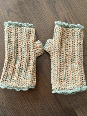 Handvirkade handledsvärmare - Ett par virkade handledsvärmare i en mönstrad design. De har en ribbad struktur. Grön/beige melerad färg med dekorativ kant.  Ca 18 cm långa. Fint skick, knappt använda. Katt i hemmet.