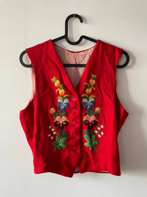 Röd broderad väst med blommor - Unik röd väst med färgglada blombroderier framtill. Västen har V-ringning, knäppning med fem knappar och är ärmlös. Materialet känns som bomull eller ullmix och insidan är ljusrosa. 