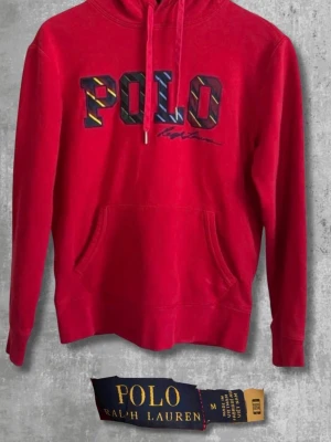 Röd hoodie från Polo Ralph Lauren - Riktigt snygg röd hoodie från Polo Ralph Lauren med stor POLO-logga i färgglada bokstäver på bröstet och signaturbrodyr under. Klassisk känguruficka framtill, justerbar huva med dragsko och mjukt bomullsmaterial. Perfekt för en avslappnad streetwear-look.
