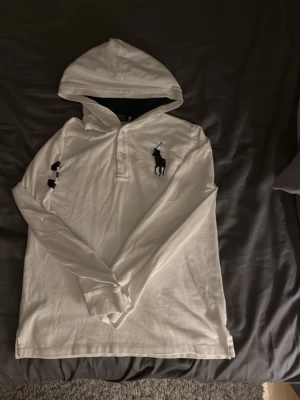 Vit hoodie från Polo Ralph Lauren - Snygg vit hoodie från Polo Ralph Lauren med klassisk marinblå logga broderad på bröstet. Hoodien har långa ärmar, knappar vid halsen och är gjord i ett mjukt bomullstyg. Perfekt för en clean och stilren look.