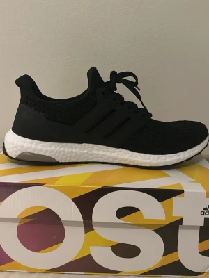 Adidas Ultra Boost svarta sneakers - Svarta Adidas Ultra Boost sneakers med stickad ovandel och vita Boost-sulor. Skorna har klassiska tre ränder på sidan, svart hälkappa och texten 'ultra boost' på hälen. Låg modell med snörning och bekväm dämpning, perfekt för en sportig look.