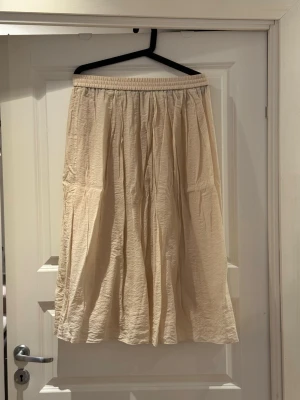 Beige långkjol med resårmidja - Luftig beige långkjol med resår i midjan och lätt rynkad struktur. Kjolen har en avslappnad passform och är tillverkad i ett mjukt, tunt tyg som ger ett flowy intryck. Perfekt för dig som gillar en enkel och stilren look.