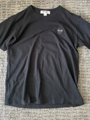 Svart Comme des Garçons PLAY t-shirt - Svart t-shirt från Comme des Garçons PLAY med det ikoniska hjärte-logotypen på bröstet. Klassisk passform, rund hals och korta ärmar. Tillverkad i mjuk bomull som känns skön mot huden. Perfekt för en clean och stilren look.