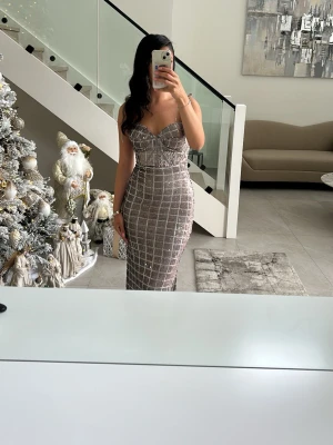 Glittrig balklänning  - Säljer en elegant långklänning från Kosovo med rutigt mönster och glittriga detaljer. Klänningen har en v-ringad front, smala axelband och figurnära passform. Perfekt för dig som vill sticka ut med en lyxig look. Materialet känns som syntet och klänningen har en slits bak.