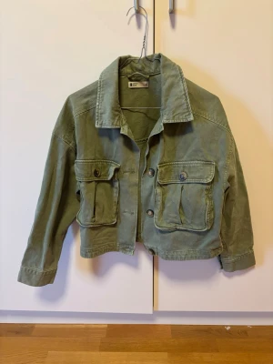 Grön cropped jeansjacka med fickor S - Snygg grön cropped jeansjacka med två stora bröstfickor med lock och knapp. Klassisk krage och raka ärmar. Jackan har en avslappnad passform och cool vintagekänsla, perfekt för att lyfta din outfit.