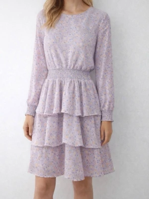 Ljuslila volangklänning med blommor - Söt ljuslila klänning med småblommigt mönster och volanger i två lager. Klänningen har långärmade ärmar med smock vid handlederna och midjan, samt rund halsringning. Perfekt för vår och sommar! Storlek 15 år  170