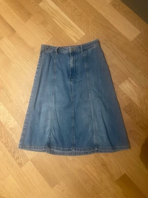 Tommy Hilfiger blå  jeanskjol i A-linje stl 36 - Säljer en klassisk blå jeanskjol i A-linjeform med hög midja och bälteshällor. Kjolen har en knapp och dragkedja framtill och är tillverkad i ett mjukt denimtyg. Perfekt att styla med sneakers eller boots för en chill look.