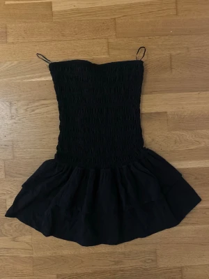 Svart bandeau jumpsuit med volang - Säljer en svart bandeau jumpsuit med smockad överdel och volangdetalj vid midjan. Kort modell med avslappnad passform och mjukt tyg som känns skönt mot huden. Perfekt för dig som vill ha en enkel men snygg look.
