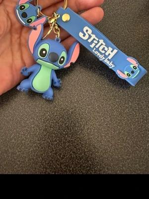Stitch nyckelring med blå rem - Nyckelring med en söt Stitch-figur i blått och grönt, samt en blå silikonrem med texten 'Stitch Lovely Baby' och små Stitch-motiv. Perfekt att hänga på väskan eller nycklarna för en lekfull touch.