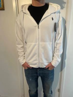 Polo Ralph Lauren hoodie   - Snygg vit hoodie från Polo Ralph Lauren med dragkedja framtill, två stora fickor och klassisk huva med dragsko. Ikoniska lilla loggan broderad på bröstet. Tillverkad i mjuk bomullsblandning som känns skön mot huden. Perfekt för en clean och stilren look.